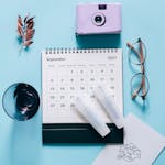 Top view of a calendar with a camera, glasses, and leaves on a pastel blue background.
