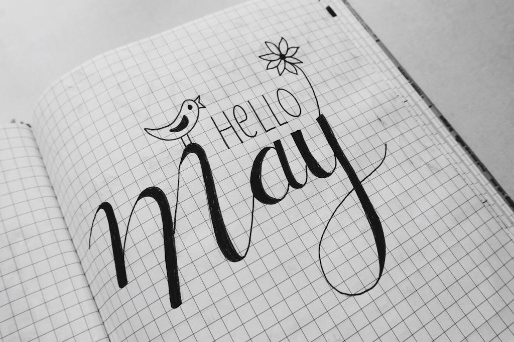 Black and white artistic calligraphy of 'Hello May' in a notebook with flower and bird doodles.